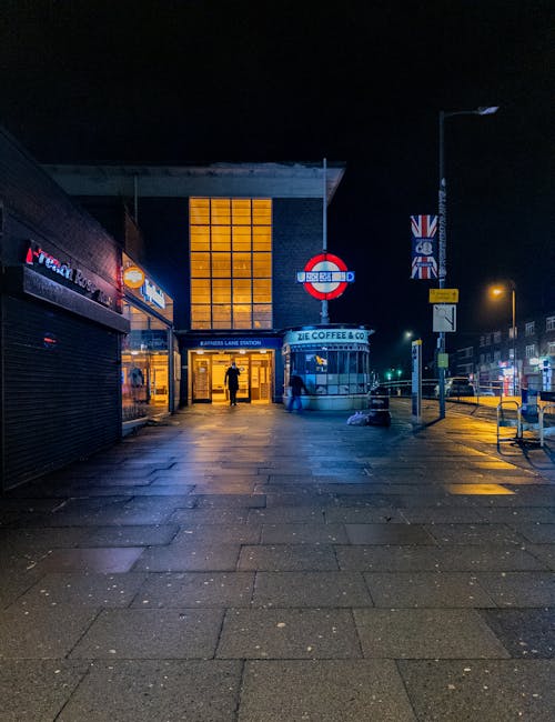 A nighttime view of the entrance to Wembley Park Underground Station, illuminated by warm orange lighting from the station's glass façade and adjacent streetlights. The station's signage is visible, 