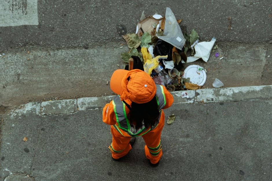 A person’s hand placing paper waste into a white recycling bin labeled 'RECYCLE' for waste segregation during domestic cleaning. The bin is situated on a wooden surface in a well-lit room. Nearby, t