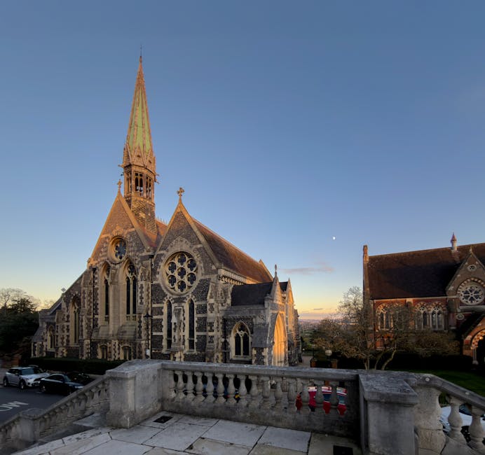 A detailed view of Headstone Manor's stone terrace overlooking a historic church with a tall, pointed spire. The church features intricate gothic architecture with stone walls, large stained-glass win