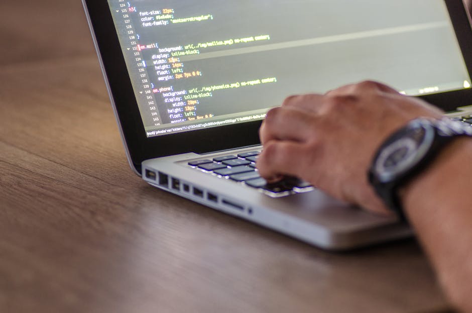 A person using a laptop placed on a wooden desk, with a visible screen displaying programming code. The individual is wearing a wristwatch and is typing on the keyboard, suggesting a focus on digital 